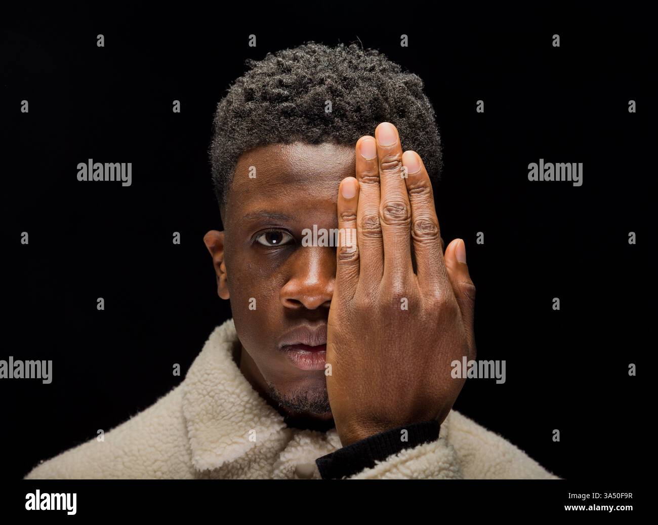 Portrait en studio d'un homme noir aux cheveux bouclés courts, couvrant partiellement son visage avec ses mains sur un fond sombre. La composition sombre met l'accent sur le calme, le charisme et une forte présence personnelle. Banque D'Images
