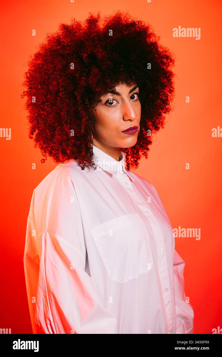Portrait d'une jeune femme hispanique aux cheveux afro foncés, portant une chemise blanche, sur un fond rouge vif. La pose transmet la confiance avec un look sans émotion et avant-gardiste, idéal pour une utilisation éditoriale et de campagne. Un portrait chic et contemporain de la tête et des épaules qui met en valeur l'individualité et le style. Banque D'Images