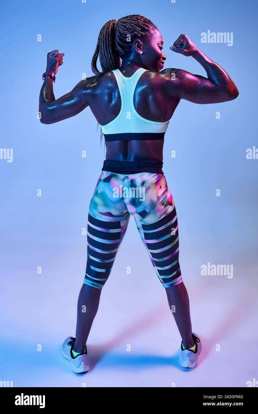 Athlète afro-américaine se tient sur le côté et montre ses biceps sur un fond bleu. Sa pose forte et athlétique met l'accent sur la forme physique, la force et la détermination. Parfait pour le sport, les femmes en fitness et les campagnes de santé. Banque D'Images
