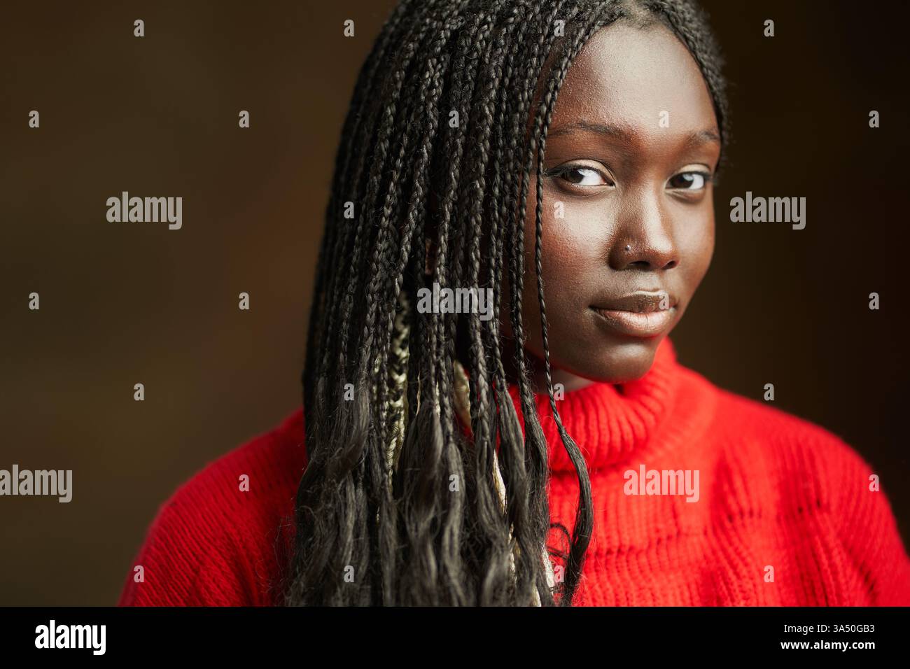 Portrait d'une jeune femme afro-américaine avec des tresses afro portant un haut à manches longues crocheté rouge dans un studio avec un fond brun. Elle regarde la caméra avec une expression calme et tranquille, présentant une beauté naturelle confiante. Idéal pour la mode, la beauté ou les portraits. Banque D'Images