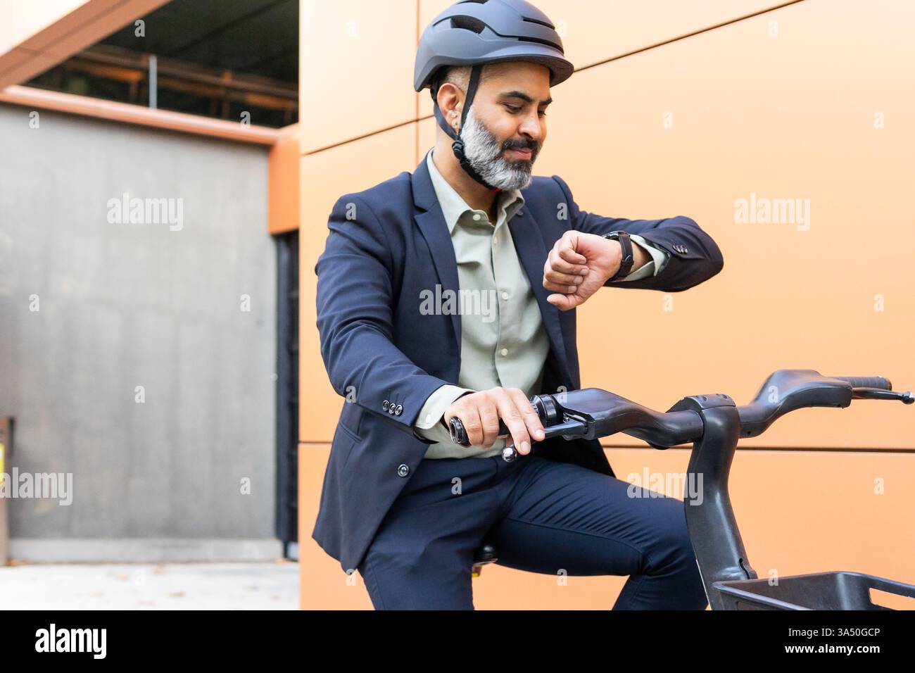 Homme indien en tenue d'affaires assis sur un vélo électrique dans un cadre urbain, vérifiant sa montre. La scène met en avant la mobilité intelligente et les déplacements écologiques. Convient à la vie urbaine, aux transports et aux thèmes technologiques. Banque D'Images