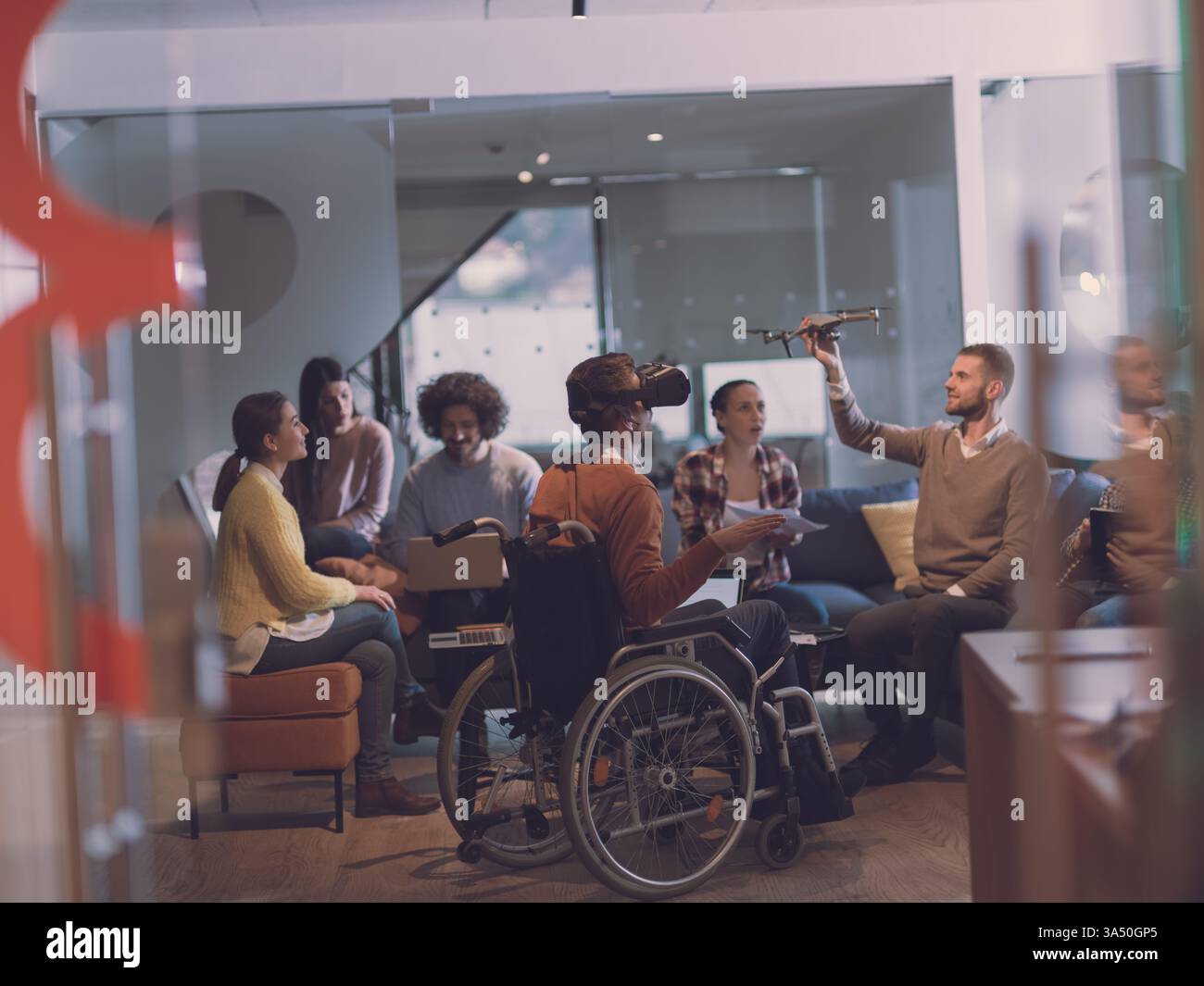 Un homme d'affaires handicapé en fauteuil roulant participe à une réunion d'équipe dans un bureau de coworking à espace ouvert moderne. Il s’engage avec des lunettes de réalité virtuelle et une assistance par drone, mettant en avant l’accessibilité et l’innovation. Une image idéale pour la technologie inclusive, la culture des startups et les lieux de travail futurs. Banque D'Images