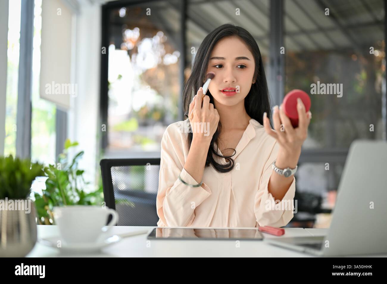 Attrayante femme d'affaires asiatique millénaire appliquant le blush à son bureau. Ce moment de beauté et de mode capture le style professionnel et le toilettage personnel sur le lieu de travail. Idéal pour les campagnes sur la beauté du lieu de travail, la mode et l'entrepreneuriat féminin. Banque D'Images