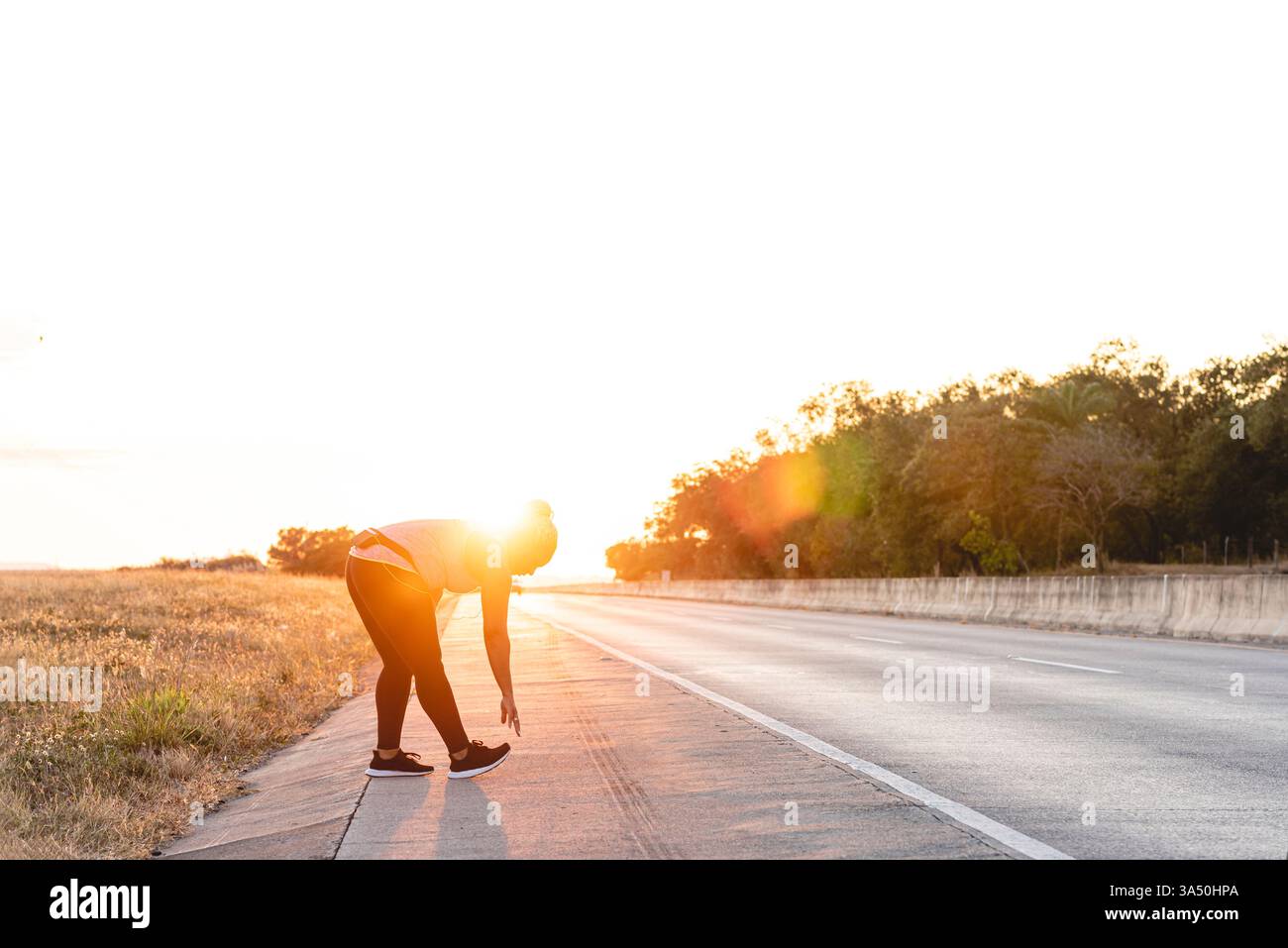 Femme afro-latina en vêtements actifs faisant des exercices d'étirement sur le bord de la route au coucher du soleil Banque D'Images