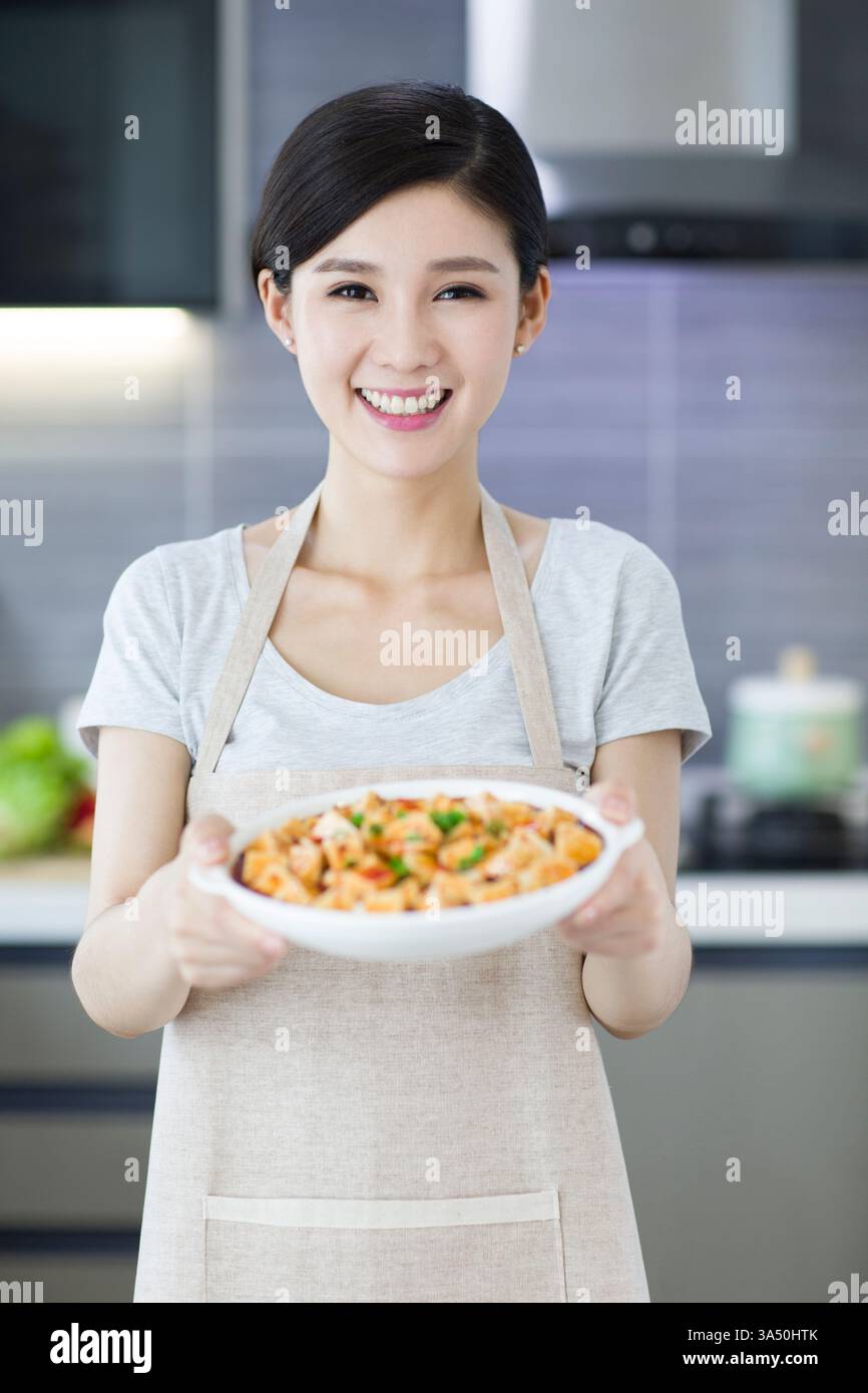 Heureuse jeune femme chinoise servant de la nourriture dans une cuisine. Le moment met en lumière la cuisine, la vie domestique et l'hospitalité. Convient pour les recettes, la vie à la maison et les thèmes du service alimentaire. Banque D'Images