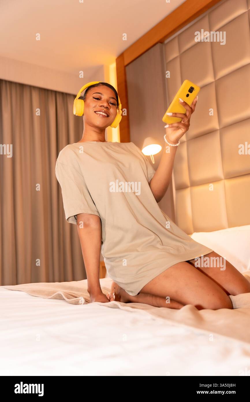 Portrait d'une femme noire souriante écoutant de la musique sur un lit avec des écouteurs. Le cadre intérieur de la chambre transmet un style de vie détendu, le confort et le style personnel. Idéal pour la musique, la vie à la maison et les campagnes de mode. Banque D'Images