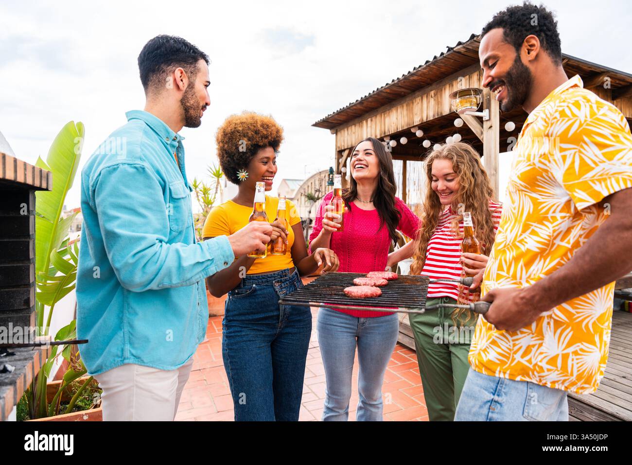 Groupe d'amis joyeux et diversifiés debout ensemble et buvant de la bière à un barbecue sur le toit pendant la journée. Cette scène sociale animée capture l'ensemble, la célébration et la vie urbaine. Idéal pour les visuels de style de vie, de fête et de campagne estivale. Banque D'Images