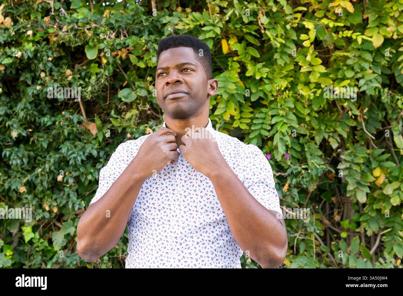 Portrait extérieur d'un homme noir touchant sa chemise, debout parmi des plantes luxuriantes à la lumière du jour. Cette image de style de vie convient à la mode, au style personnel ou aux contextes de bien-être extérieur. Banque D'Images