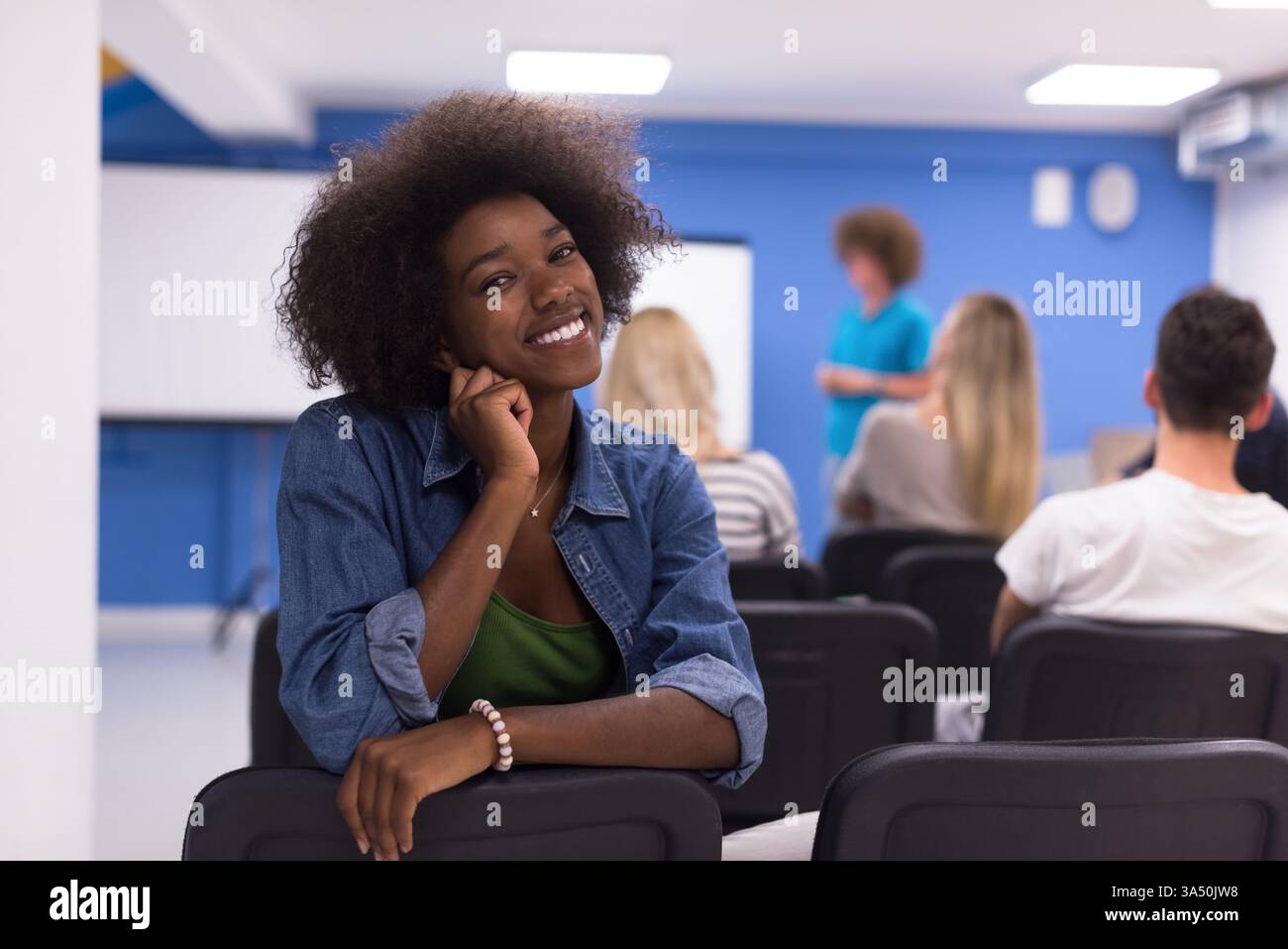Joyeuse femme noire avec un afro est assise à l'envers dans une salle de conférence, souriant à côté de collègues pendant une réunion. Idéal pour la communication d'entreprise, le travail d'équipe et la diversité sur le lieu de travail dans les environnements de bureau modernes. Banque D'Images