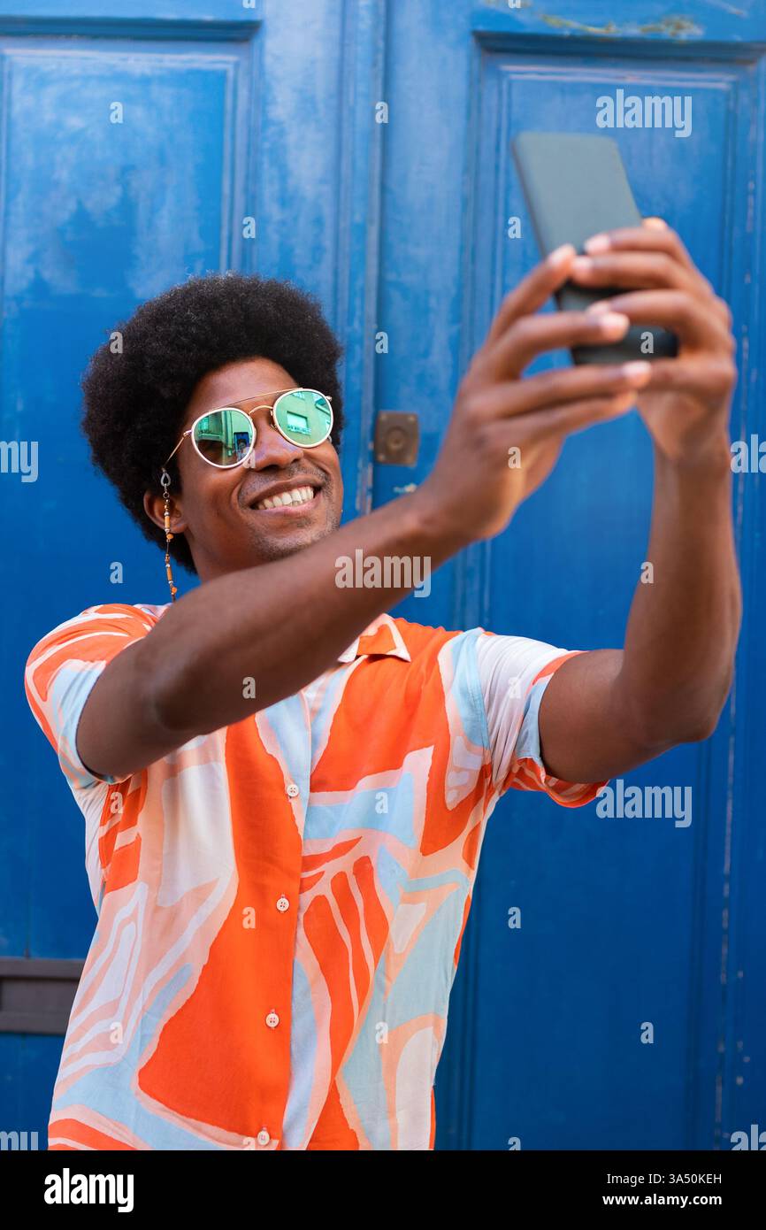 Homme afro-américain souriant avec un afro prend un selfie sur un smartphone sur un fond de rue bleu. Ce portrait extérieur optimiste convient parfaitement aux thèmes de mode de vie, de médias sociaux et de mode urbaine. Idéal pour l'image de marque personnelle et les visuels de style urbain contemporain. Banque D'Images