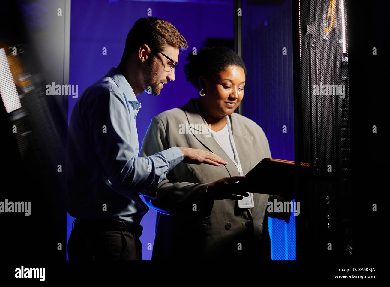 Portrait de taille haute de deux administrateurs système utilisant un ordinateur dans une salle de serveur sombre éclairée par un néon, avec espace de copie pour le texte. L'image axée sur la technologie met l'accent sur les centres de données, la mise en réseau et la collaboration. Banque D'Images