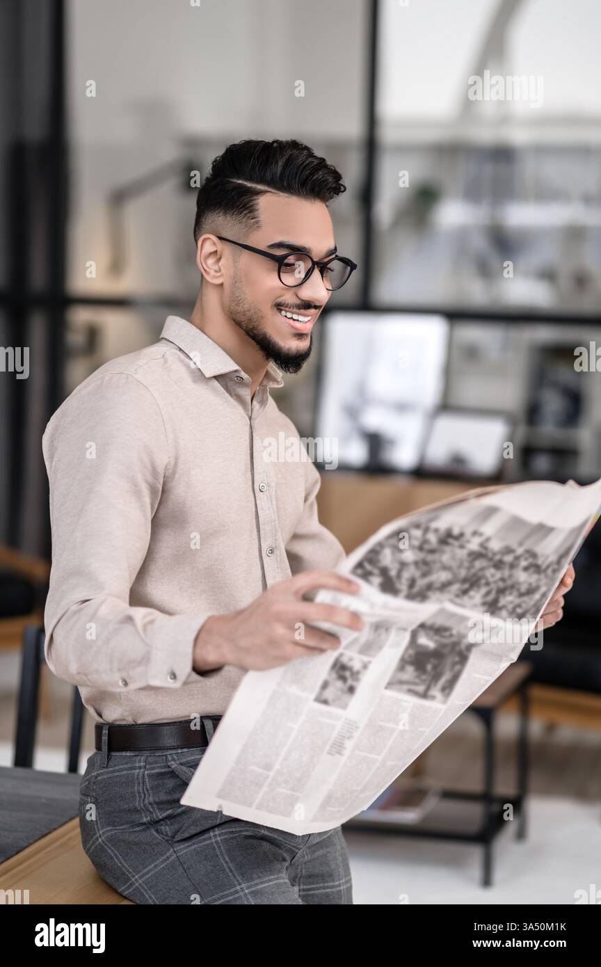 Joyeux jeune homme barbu dans des verres regarde un journal à l'intérieur, debout et appuyé sur une table pendant la journée. La scène transmet des informations et une ambiance professionnelle confiante et élégante. Idéal pour les affaires, les actualités et le contenu de style de vie des travailleurs indépendants. Banque D'Images