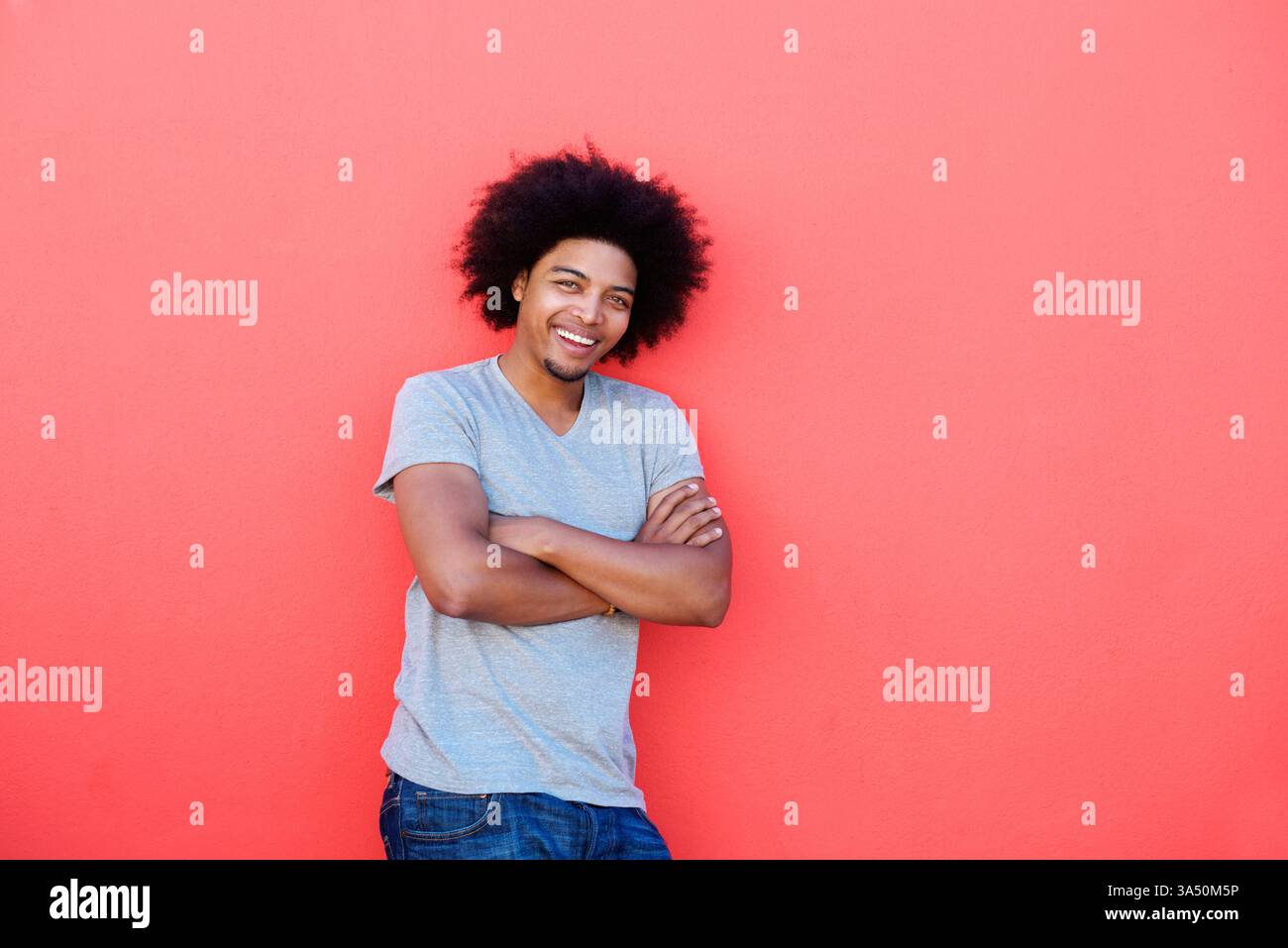 Portrait d'un jeune homme confiant avec un afro souriant avec les bras croisés sur un fond rouge audacieux. Un portrait de style de vie saisissant adapté à la mode, à la marque personnelle et aux images d'humeur positive. Banque D'Images