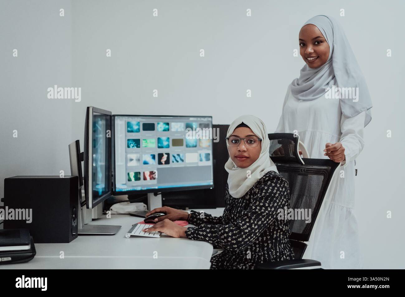 Deux jeunes femmes musulmanes afro-américaines en foulards travaillent ensemble dans un bureau lumineux et moderne avec un grand écran. Cette scène met en lumière le travail d’équipe et la collaboration professionnelle dans un milieu de travail contemporain. Banque D'Images
