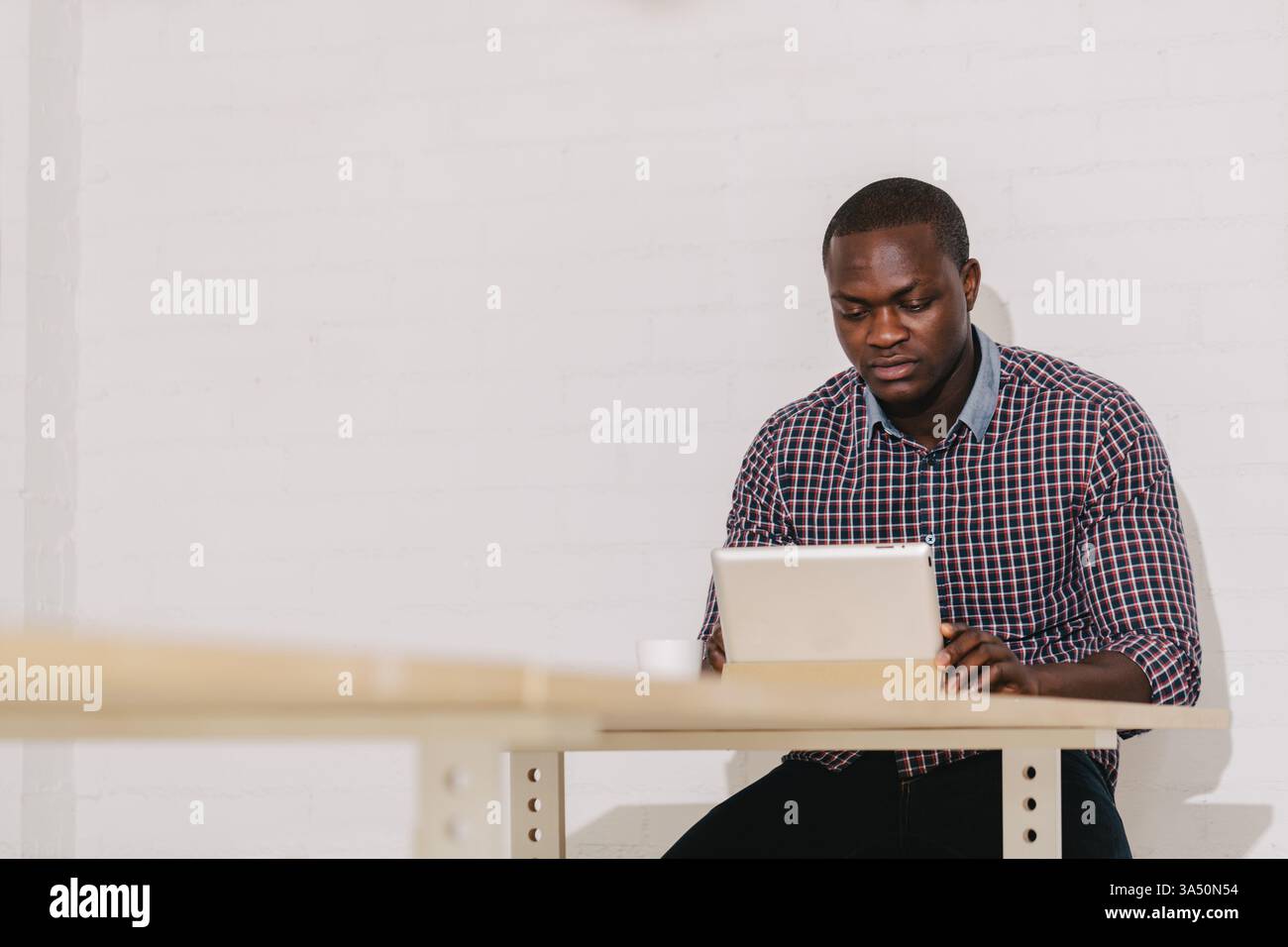Homme noir chauve sérieux travaillant sur un ordinateur portable assis à une table dans un bureau. Une scène professionnelle ciblée adaptée aux thèmes des affaires, de la technologie et du travail à distance. Idéal pour les campagnes de communication d'entreprise, de productivité et d'environnement de travail moderne. Banque D'Images