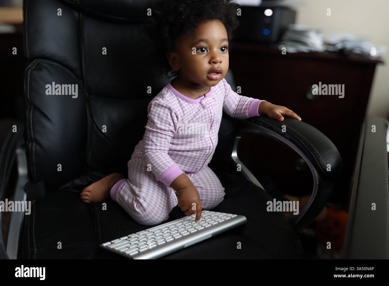 Black Baby Girl joue avec un clavier sans fil tout en étant assis sur une chaise de bureau en cuir à la maison. Un moment chaleureux et convivial pour les tout-petits, pour des visuels familiaux et technologiques. Banque D'Images