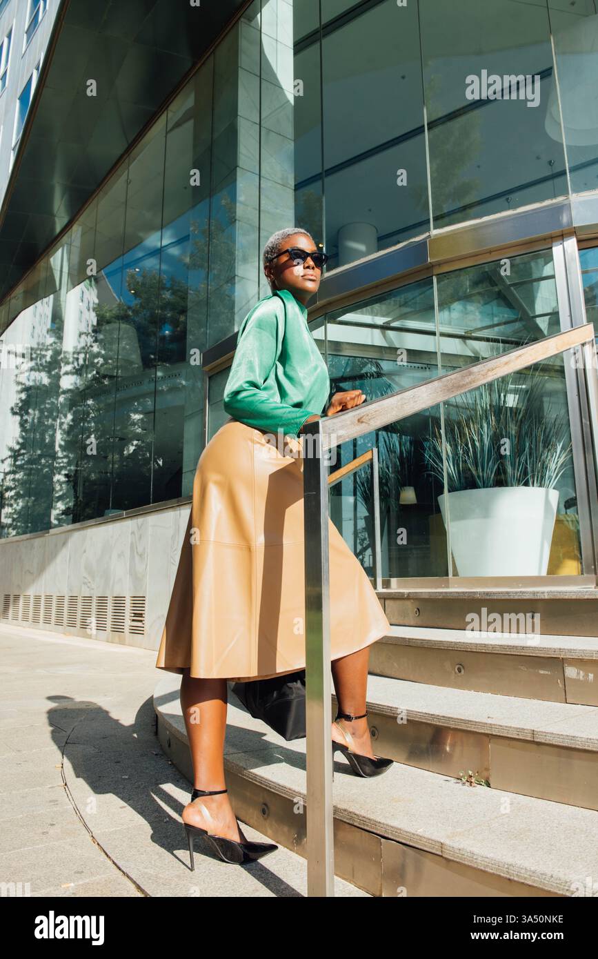 Femme d'affaires noire portant des lunettes de soleil monte les escaliers à l'intérieur d'un bâtiment en verre. L'image élégante et confiante convient à la mode urbaine, au leadership et aux campagnes de style de vie d'entreprise. Idéal pour les caractéristiques sur les femmes dans les affaires et l'architecture de la ville. Banque D'Images