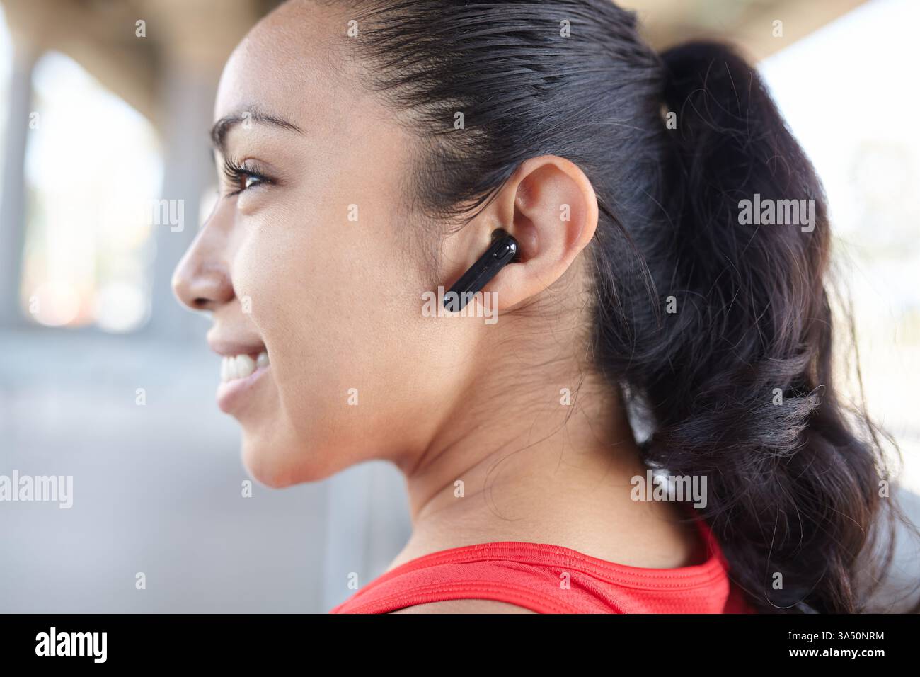 Une femme hispanique optimiste en vêtements de fitness se tient à l'extérieur sous un pont après une séance d'entraînement, portant des écouteurs sans fil. Elle sourit en toute confiance, soulignant un style de vie actif lorsqu'elle écoute de la musique sur son appareil. Banque D'Images