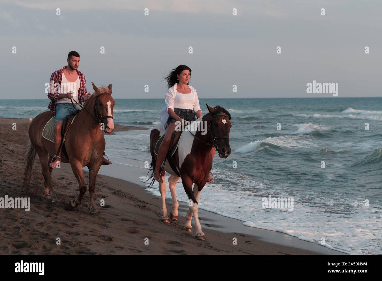 Deux cavaliers, une femme mixte en haut blanc et en jeans courts, et son petit ami caucasien, montent à cheval le long d'une plage au coucher du soleil. La scène capture une lumière dorée chaude, des vues sur l'océan et une ambiance romantique idéale pour les voyages, les couples et les loisirs en plein air. Idéal pour les campagnes lifestyle mettant en vedette la plage, la romance et les moments de vacances. Banque D'Images