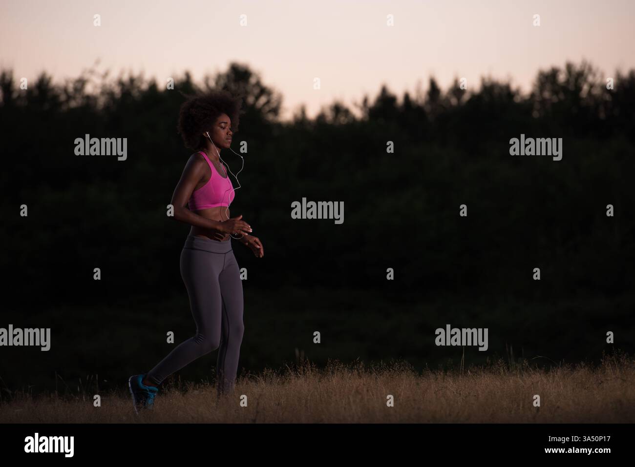 Jeune femme afro-américaine faisant du jogging en plein air dans la nature pendant une nuit d'été, portant des écouteurs. Cette scène de remise en forme transmet de l'énergie, de la vitalité et de l'entraînement en plein air. Convient pour les campagnes sur la course à pied, les entraînements cardio et une vie saine. Banque D'Images