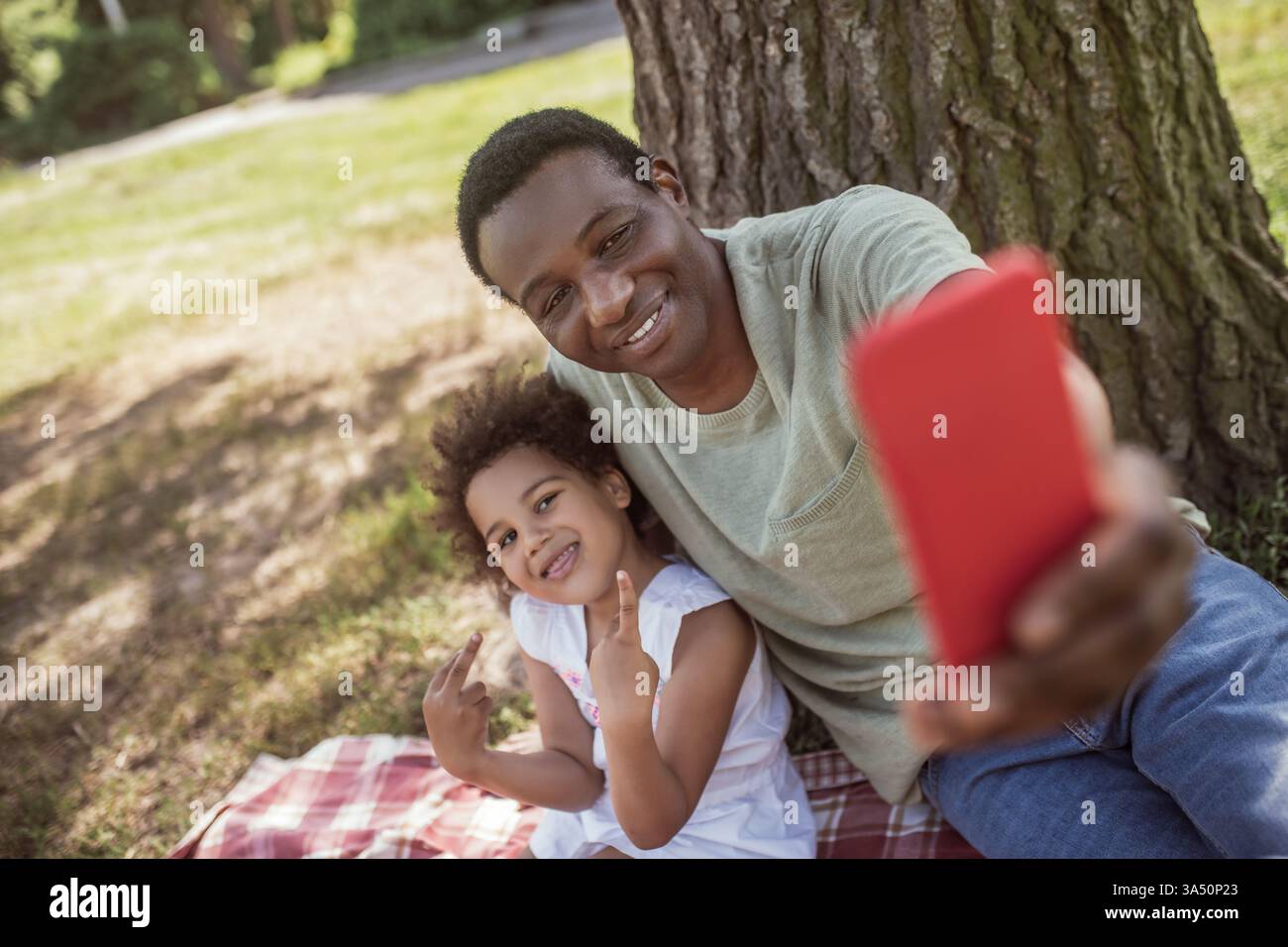 Un homme souriant à la peau foncée prend un selfie avec sa jeune fille dans un parc. Un moment familial chaleureux idéal pour les campagnes parentales, paternelles et lifestyle. Banque D'Images