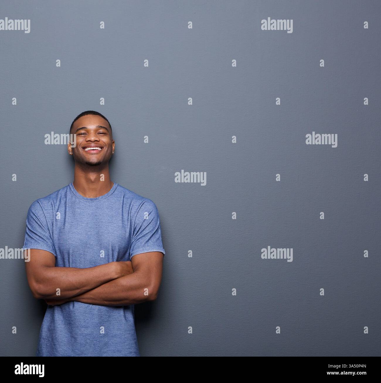 Homme afro-américain souriant avec les bras pliés se tient sur un fond gris. Ce portrait de style de vie confiant capture une ambiance moderne et accessible, idéale pour la marque personnelle et les visuels marketing. Convient aux campagnes commerciales, technologiques et de style de vie contemporain. Banque D'Images