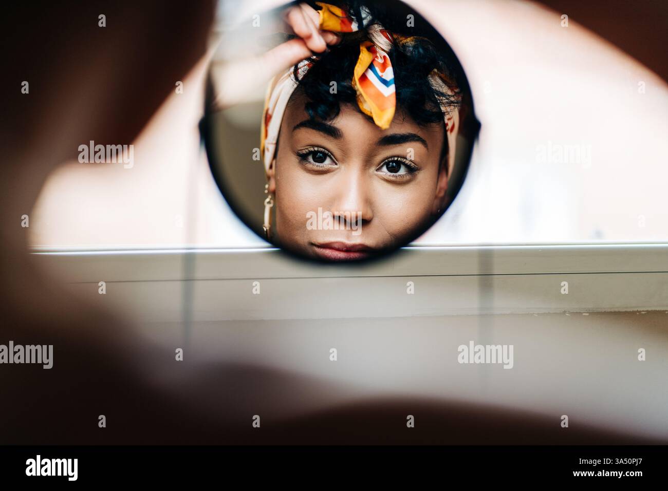 Femme noire à la maison, vue dans un miroir rond alors qu'elle met sur un bandeau. Une image de beauté et d'auto-soin accessible adaptée à la mode, au bien-être et aux thèmes de style personnel. Banque D'Images