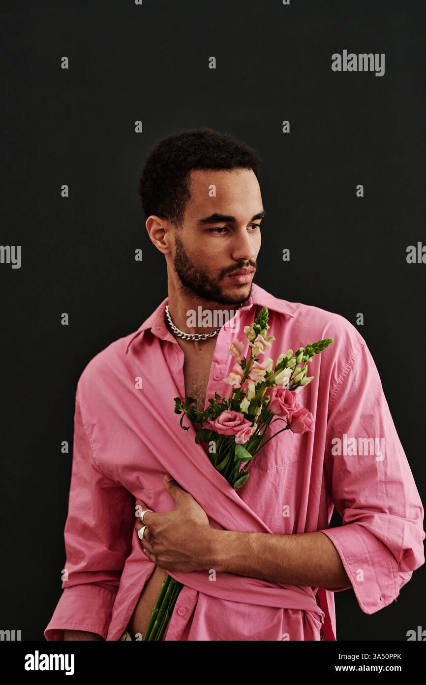 Portrait studio d'un jeune homme de race mixte posant dans une chemise rose avec des fleurs sur un fond noir. Le style vibrant et l'ambiance de la mode contemporaine conviennent à la beauté, au style de vie ou aux campagnes LGBTQ+. Idéal pour les éditoriaux de mode, les médias sociaux ou les projets de branding. Banque D'Images