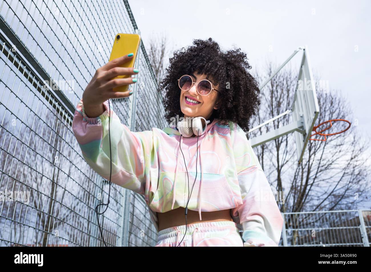 Faible angle de jeunes femmes hispaniques positives avec des lunettes de soleil de coiffure afro regardant l'écran de smartphone dans la main et prenant selfie dans le terrain de sport Banque D'Images