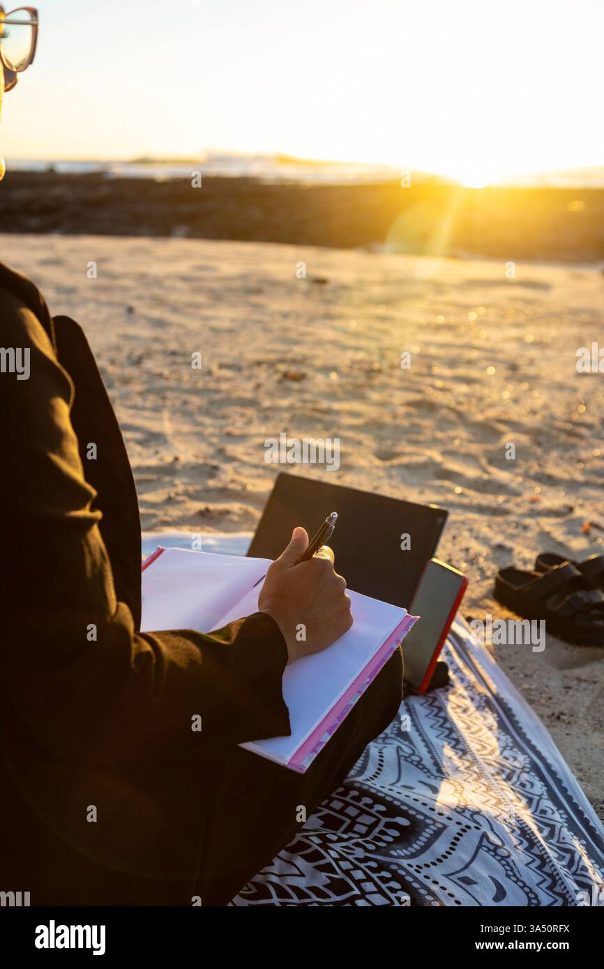 Image recadrée montre un nomade numérique non identifiable travaillant sur une tablette sur une plage de sable au coucher du soleil. Cette scène de bureau mobile met en avant le travail à distance, la connectivité et les déplacements dans un cadre côtier. Idéal pour le style de vie indépendant, le nomade numérique et les fonctionnalités de la technologie sans fil. Banque D'Images