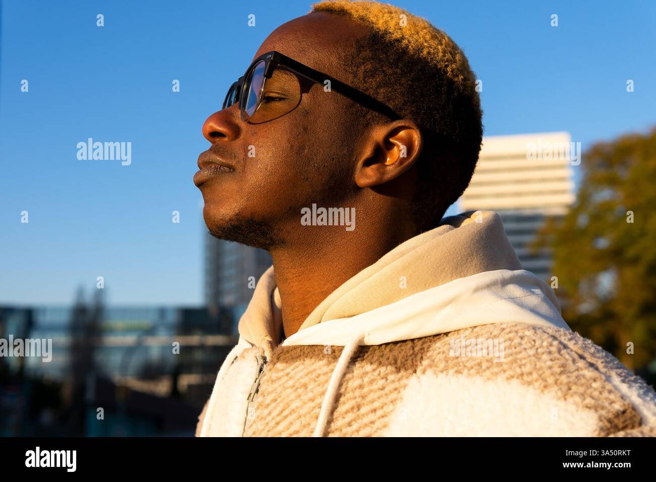 Portrait urbain réfléchi d'un homme noir debout dans une rue de la ville au coucher du soleil. Le style urbain décontracté et la lumière chaude du soir créent une ambiance contemplative, parfaite pour la photographie urbaine et de style de vie. Banque D'Images