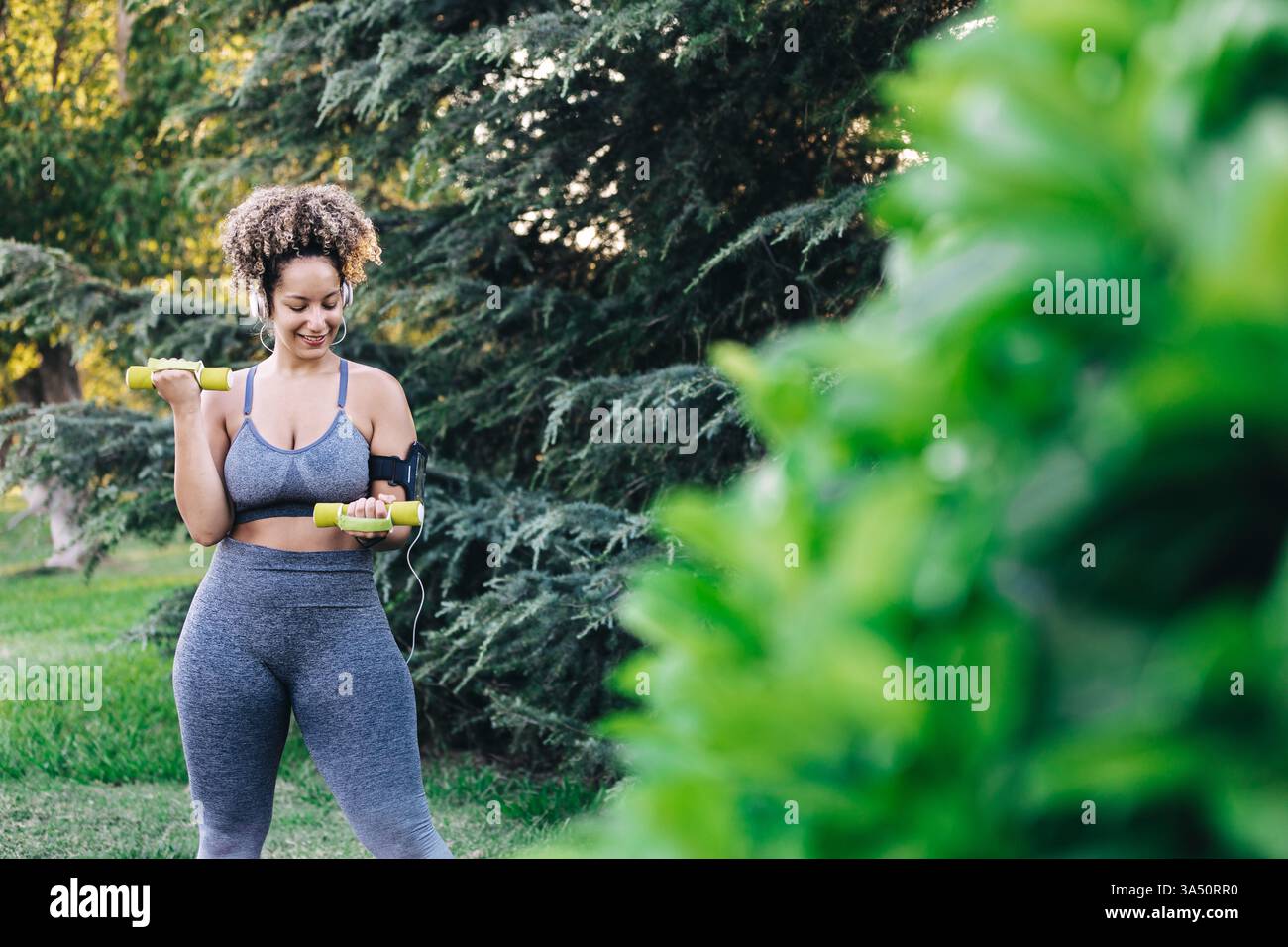 Femme hispanique dans des exercices de fitness en plein air dans un parc, soulevant des haltères. Elle porte des écouteurs et aime la musique, montrant de la motivation et de l'énergie. Idéal pour les campagnes de fitness, de bien-être et de style de vie mettant en vedette des adultes actifs. Banque D'Images