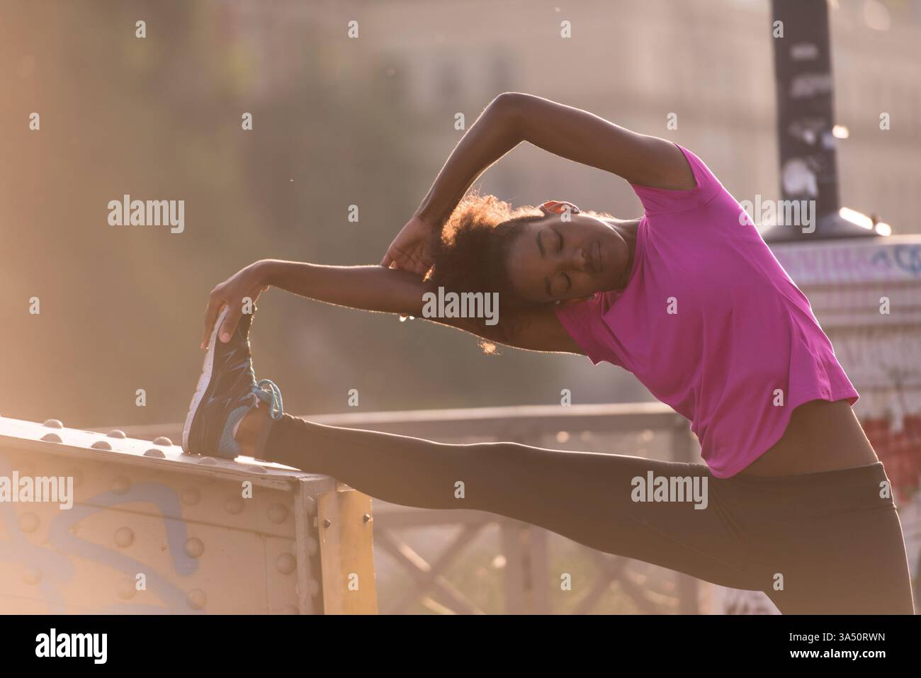 Femme afro-américaine en vêtements de sport effectue un étirement chauffant avec une jambe levée avant une course au lever du soleil. Capturé en extérieur dans un cadre urbain avec un pont et des rues de la ville, le cliché respire l'énergie et la forme physique. Parfait pour le sport, une vie saine et des campagnes de style de vie actif. Banque D'Images