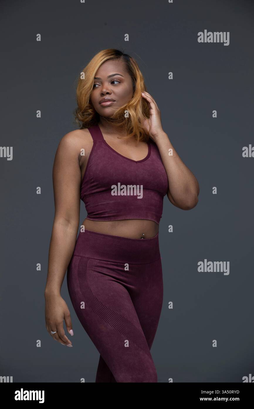 Femme noire dans un studio portant une tenue de fitness avec des jambes croisées, touchant ses cheveux sur un fond gris. Un portrait épuré et athlétique qui convient aux campagnes de bien-être, de fitness et de beauté. Idéal pour les articles sur les styles de vie actifs et les images de gymnastique. Banque D'Images