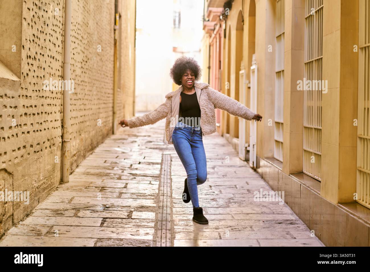Photo du corps entier d'une femme afro-américaine joyeuse dansant dans une rue résidentielle ensoleillée. Elle porte des vêtements décontractés et arbore une coiffure afro, souriant avec une ambiance insouciante et active. Parfait pour le style de vie, la danse et les visuels d'énergie positive dans les environnements urbains. Banque D'Images