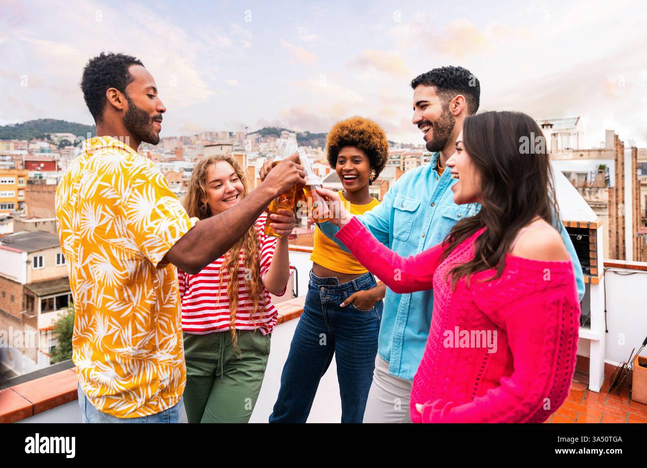 Lumineux et joyeux, un groupe diversifié d'amis porte un toast avec de la bière sur un toit-terrasse ensoleillé. Cette scène animée capture l'amitié, la convivialité et un style de vie urbain en plein air sur un balcon au-dessus de la ville. Idéal pour la vie sociale, la fête et les campagnes de style de vie estivale. Banque D'Images