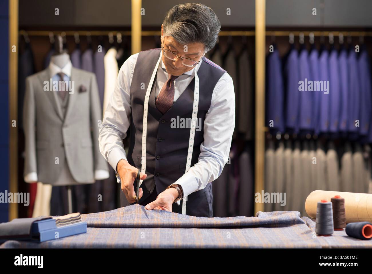 Designer de mode chinois senior se tient à une table de travail, ruban à mesurer autour du cou, coupant le tissu dans un studio de magasin de vêtements. La scène met l'accent sur l'expertise dans le design de mode, la couture et la mode pour hommes. Banque D'Images