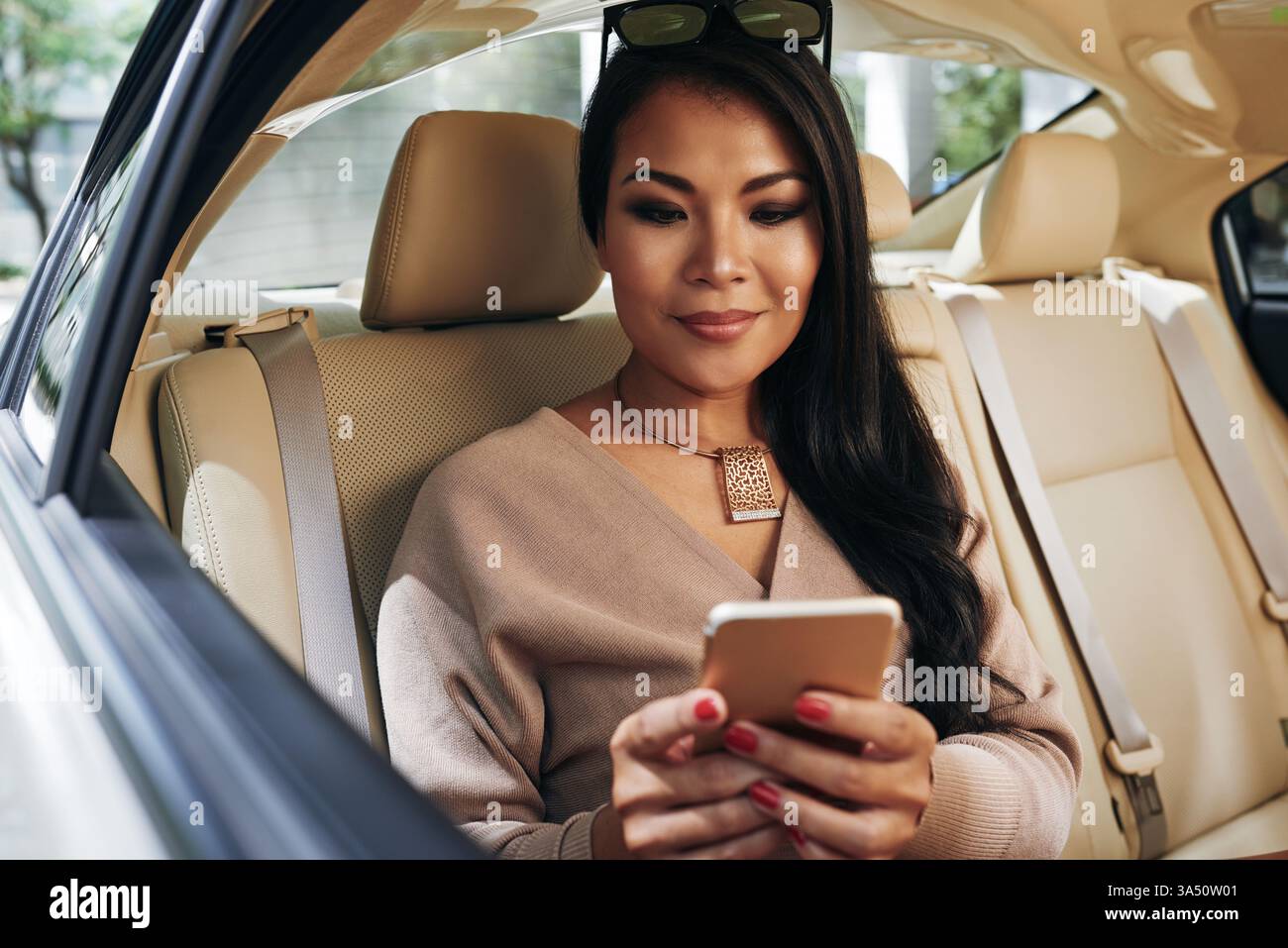 Jeune femme d'affaires asiatique est assise sur le siège arrière d'une voiture de luxe, textant sur son téléphone. Son collier ajoute une touche d'élégance lorsqu'elle travaille en déplacement. Adapté au style de vie professionnel, aux déplacements urbains et aux communications mobiles. Banque D'Images