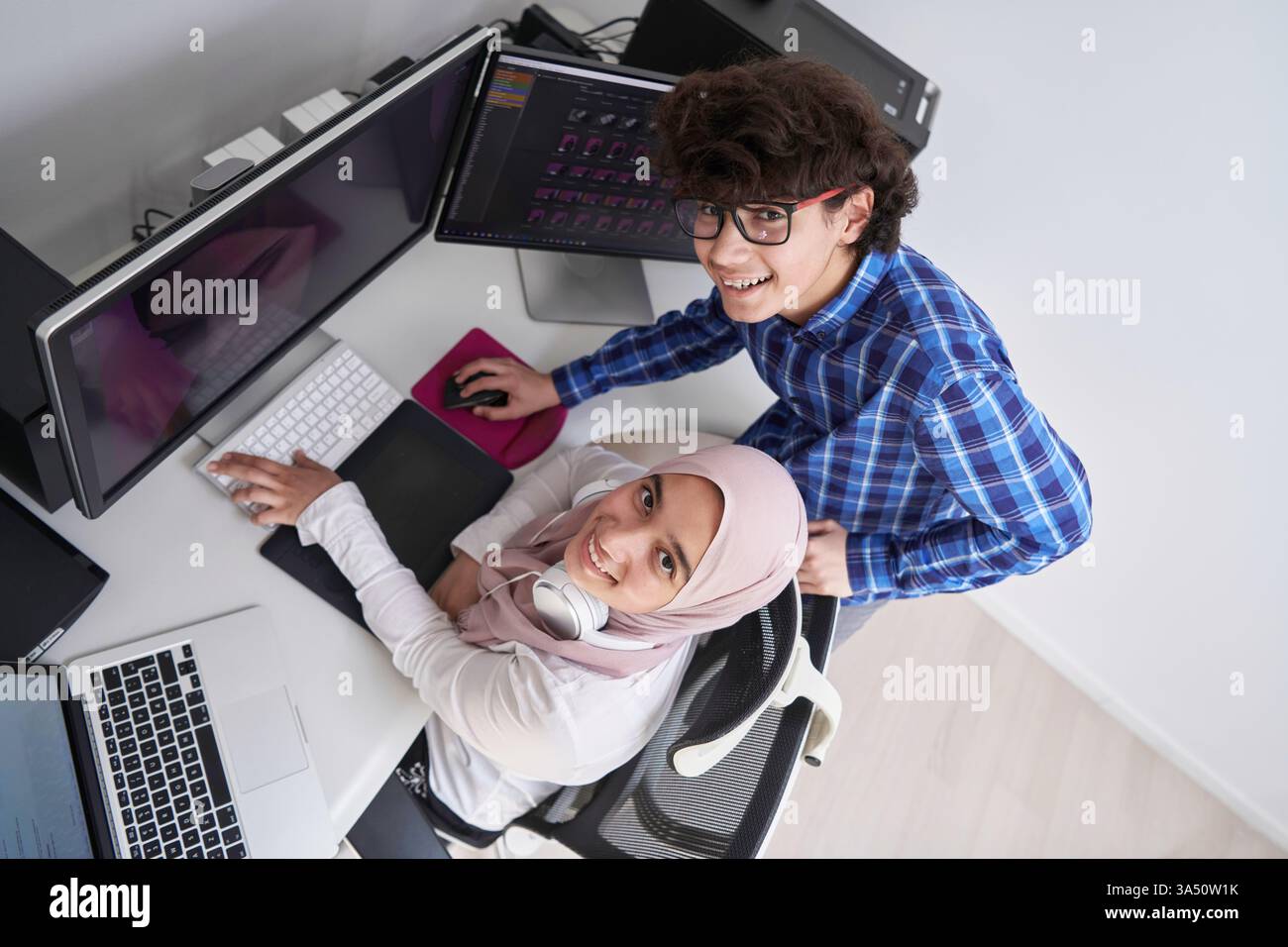 Un adolescent masculin du moyen-Orient souriant se tient à côté d'une camarade de classe musulmane alors qu'ils travaillent sur un projet scolaire sur l'ordinateur à la maison. Une scène positive et collaborative idéale pour l’éducation, la diversité et le travail d’équipe des jeunes. Banque D'Images