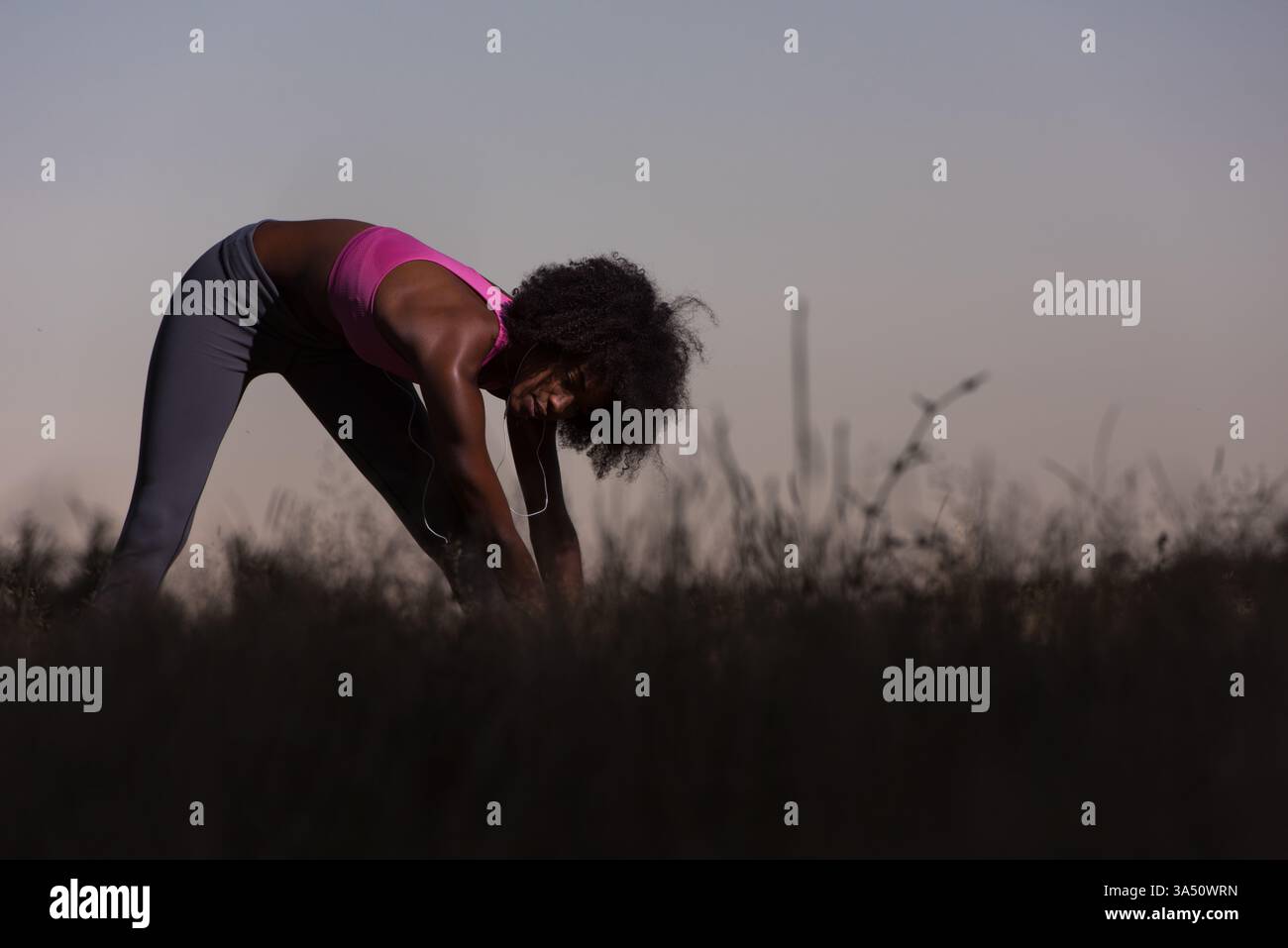 Jeune femme noire en bonne santé s'étire et se réchauffe après un jogging dans un pré pendant une soirée d'été. Cette image de fitness en plein air transmet le bien-être, la vitalité et la nature. Banque D'Images