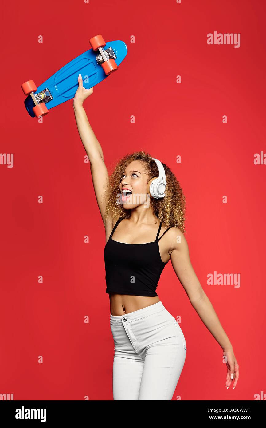 Femme hispanique joyeuse aux cheveux blonds bouclés porte des écouteurs et tient une planche à penny, posée sur un fond rouge vif. Son énergie insouciante et amoureuse de la musique ajoute une ambiance audacieuse de style urbain, idéale pour le style de vie des jeunes ou les articles de mode. Studio Shot avec une composition verticale souligne la couleur vibrante et l'ambiance moderne. Banque D'Images