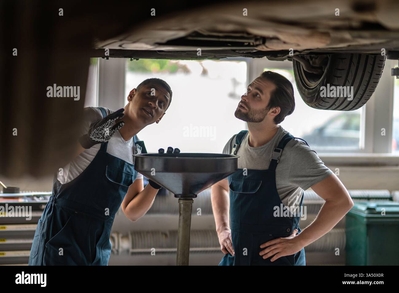 Mécaniciens noirs et caucasiens inspectant une voiture sous un véhicule soulevé dans un atelier de réparation automobile. La scène met en lumière le travail d'équipe, la diversité et le service automobile professionnel. Idéal pour l'entretien des véhicules, les ateliers et les campagnes d'entretien automobile. Banque D'Images