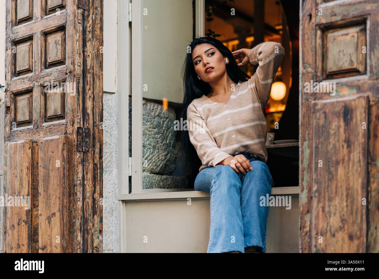 Jeune femme hispanique dans des vêtements décontractés touche ses cheveux tout en regardant loin près d'un rebord de fenêtre de café. Portrait de style de vie décontracté parfait pour la mode, la beauté ou les campagnes culturelles de café contemporain. Idéal pour le contenu des médias sociaux, le style de marque ou les blogs de style de vie en milieu urbain. Banque D'Images