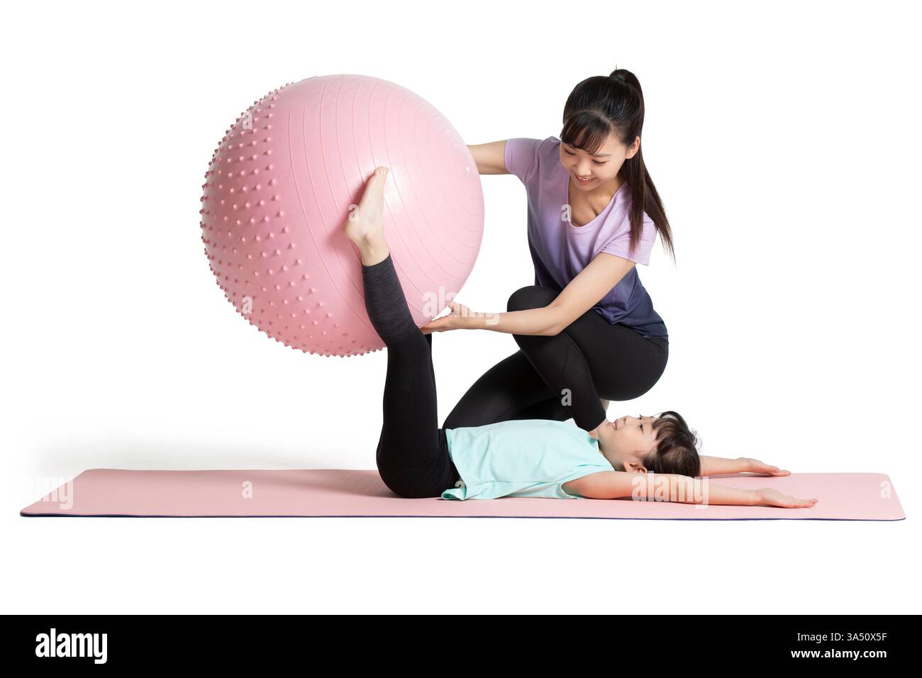 Mère et fille chinoises font de l'exercice ensemble dans un studio sur fond blanc. La scène joyeuse comprend un ballon de fitness et des conseils doux, démontrant la forme physique de la famille et un mode de vie sain. Idéal pour les activités parentales, le bien-être et les campagnes de fitness des enfants. Banque D'Images