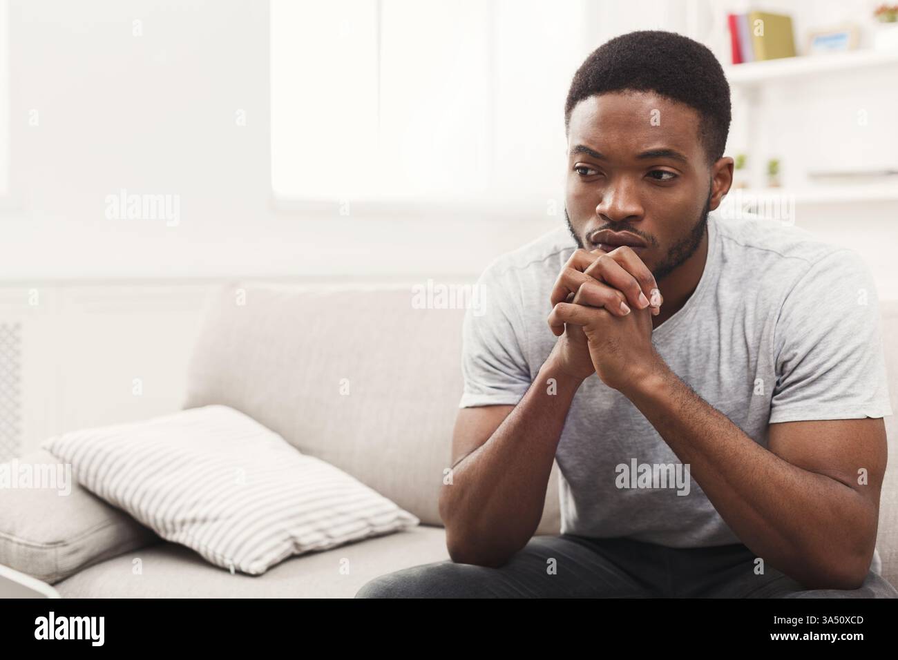 Jeune homme afro-américain assis sur un canapé à la maison avec une expression triste et réfléchie et un espace de copie. Un portrait axé sur l'humeur adapté aux sujets de santé mentale, de bien-être ou de style de vie. Idéal pour les articles, les campagnes et les publications sur les médias sociaux traitant des émotions ou de la solitude. Banque D'Images
