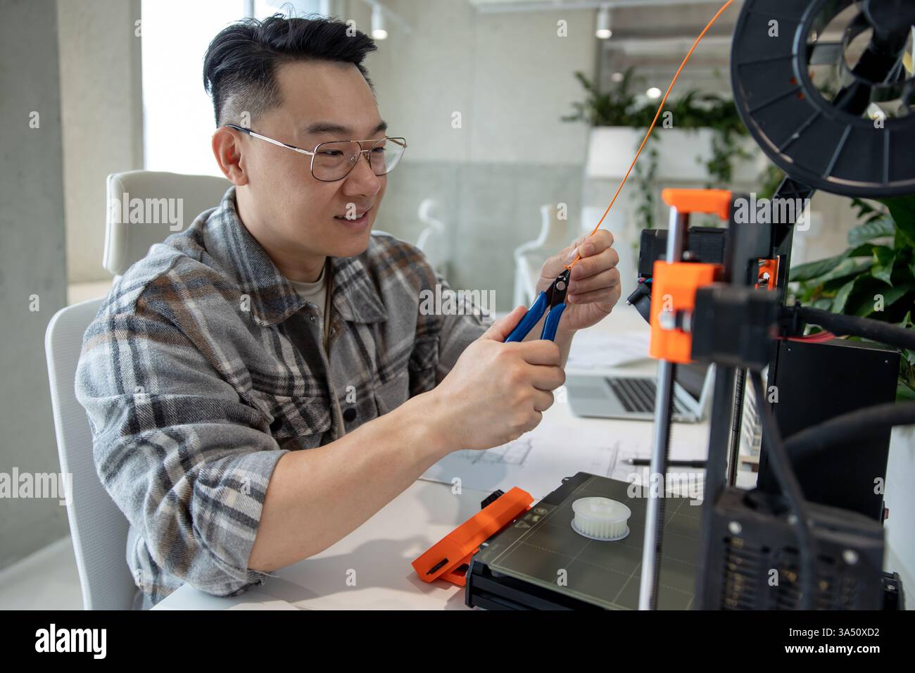 L'homme asiatique dans un bureau moderne conçoit des pièces mécaniques à l'aide d'une imprimante 3D. Cette image d'ingénierie et de technologie est idéale pour les sujets STEM, d'innovation et de fabrication. Idéal pour les articles sur la conception, le prototypage et la fabrication numérique. Banque D'Images