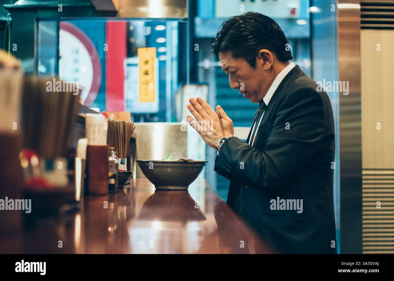 Homme d'affaires asiatique dans un restaurant est assis à une table avec des nouilles ramen. Il agrafe ses mains près de sa poitrine et tient des baguettes, ajoutant une touche contemporaine et raffinée. Idéal pour les repas d'affaires, la cuisine asiatique et les images sur le lieu de travail. Banque D'Images