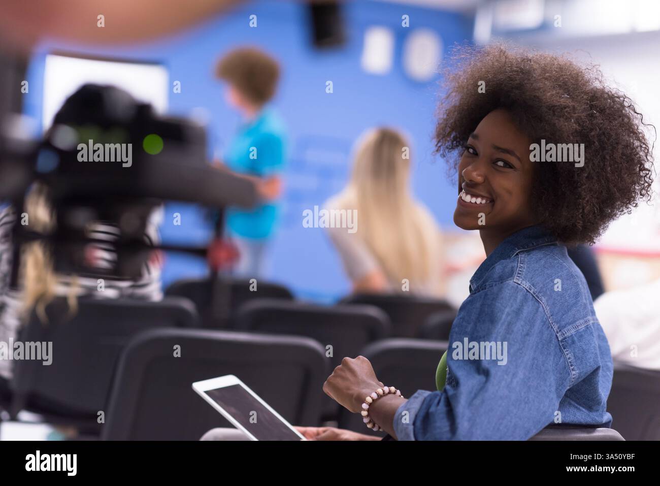 Joyeuse femme afro-américaine avec un Afro tient une tablette tout en étant assise près de collègues pendant une réunion dans une salle de conférence. La scène transmet le travail d'équipe, la technologie et une atmosphère d'affaires positive. Banque D'Images