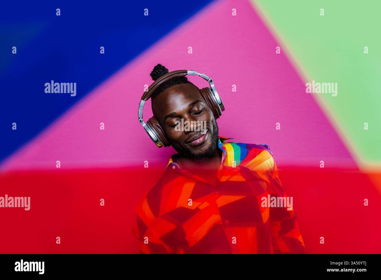 Homme afro-américain souriant portant des écouteurs se tient dans un studio avec des lumières de gel colorées. Ce portrait créatif est idéal pour la musique, les médias et les campagnes de style de vie. Banque D'Images