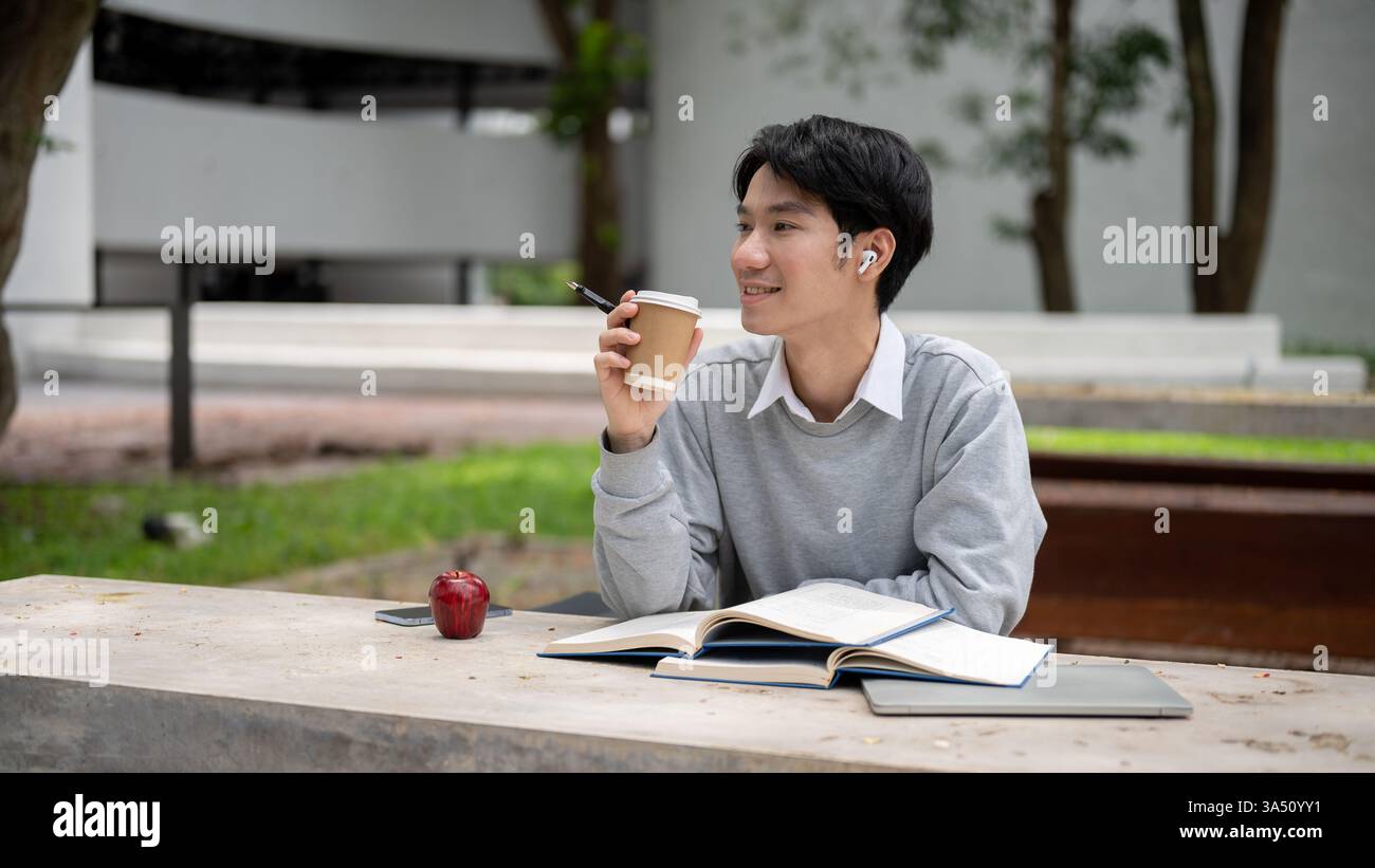 Étudiant asiatique souriant regardant loin tenant un café assis avec des livres à table sur le campus pendant la journée Banque D'Images
